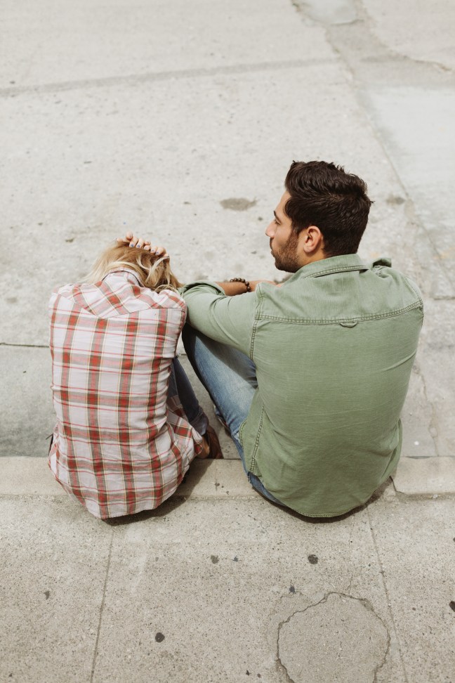 This photo shows the backs of a man and a woman sitting on a curb together.